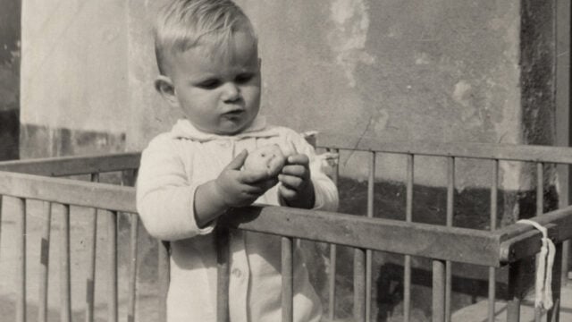 A baby with light hair stands in a wooden playpen, wearing a light-colored outfit, and holds an object—possibly a small toy or potato—while looking at it thoughtfully, evoking Boomer nostalgia for depression-era habits. The background is a rough plaster wall.