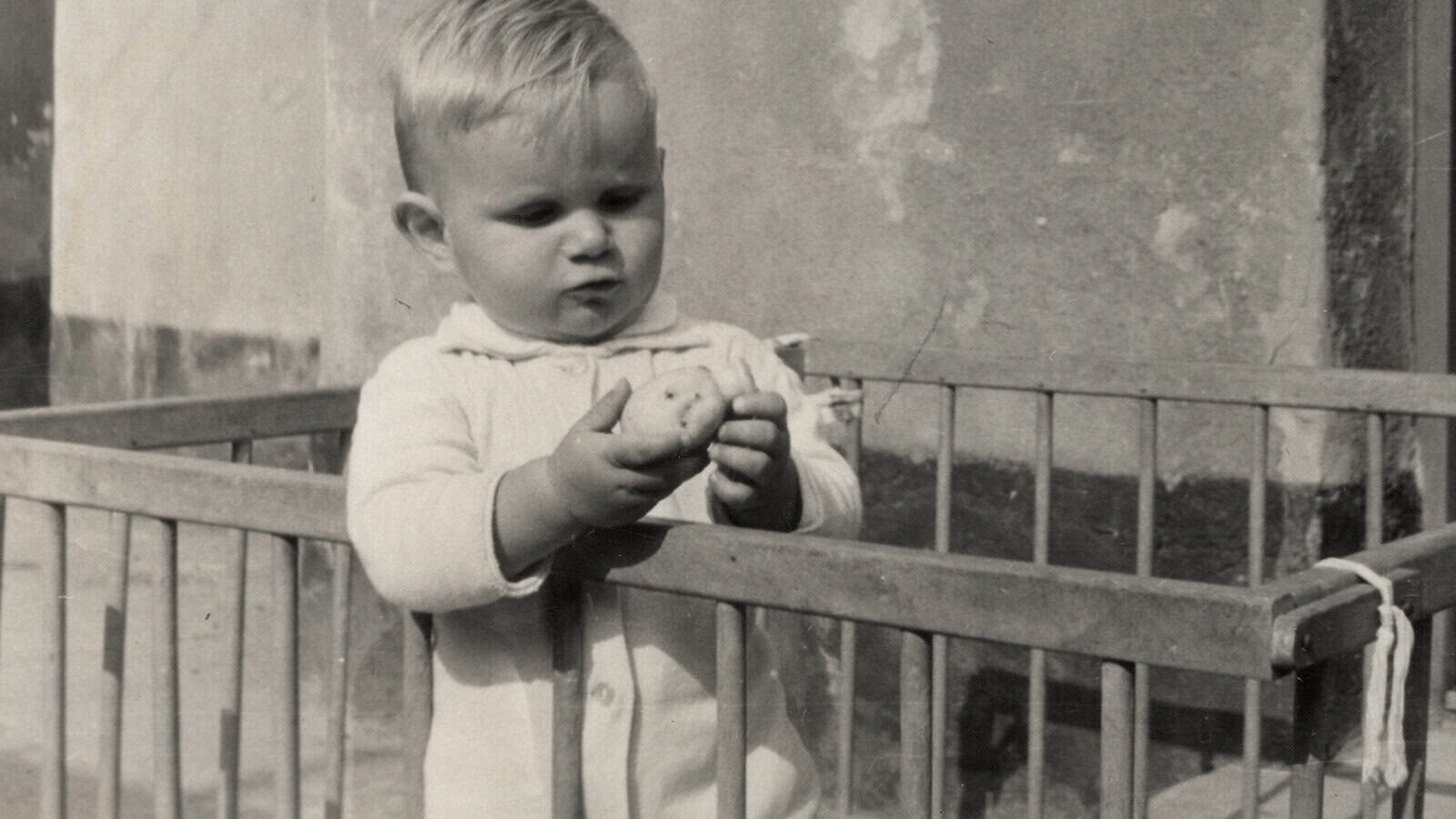 A baby with light hair stands in a wooden playpen, wearing a light-colored outfit, and holds an object—possibly a small toy or potato—while looking at it thoughtfully, evoking Boomer nostalgia for depression-era habits. The background is a rough plaster wall.
