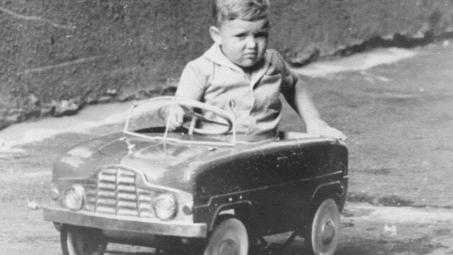 A young child in vintage clothing sits in a small pedal car, holding the steering wheel, outdoors on a paved surface. The photo is black and white, and the child looks directly at the camera with a serious expression—a reminder of an era when communication substitutes were common and many boomers learned to express themselves without always being encouraged to talk about feelings.
