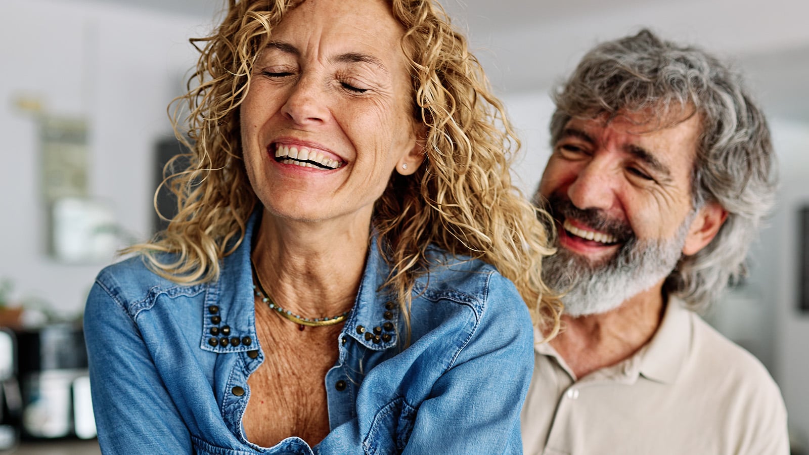 A smiling woman with curly hair in a denim shirt stands in front of a smiling man with gray hair and a beard. Both appear to be laughing together in a brightly lit indoor setting, perfectly capturing the joy and connection often seen among successful couples.