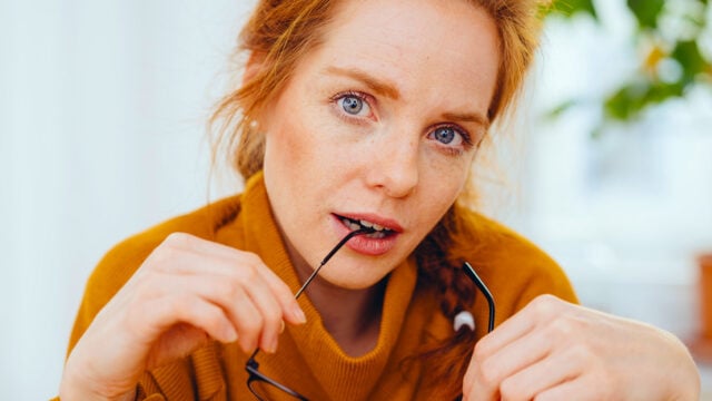 A woman with red hair and freckles in a mustard sweater holds glasses and looks thoughtfully at the camera, lightly biting the temple arm, as if contemplating the unique strengths that daughters often develop while navigating relationships with difficult mothers.