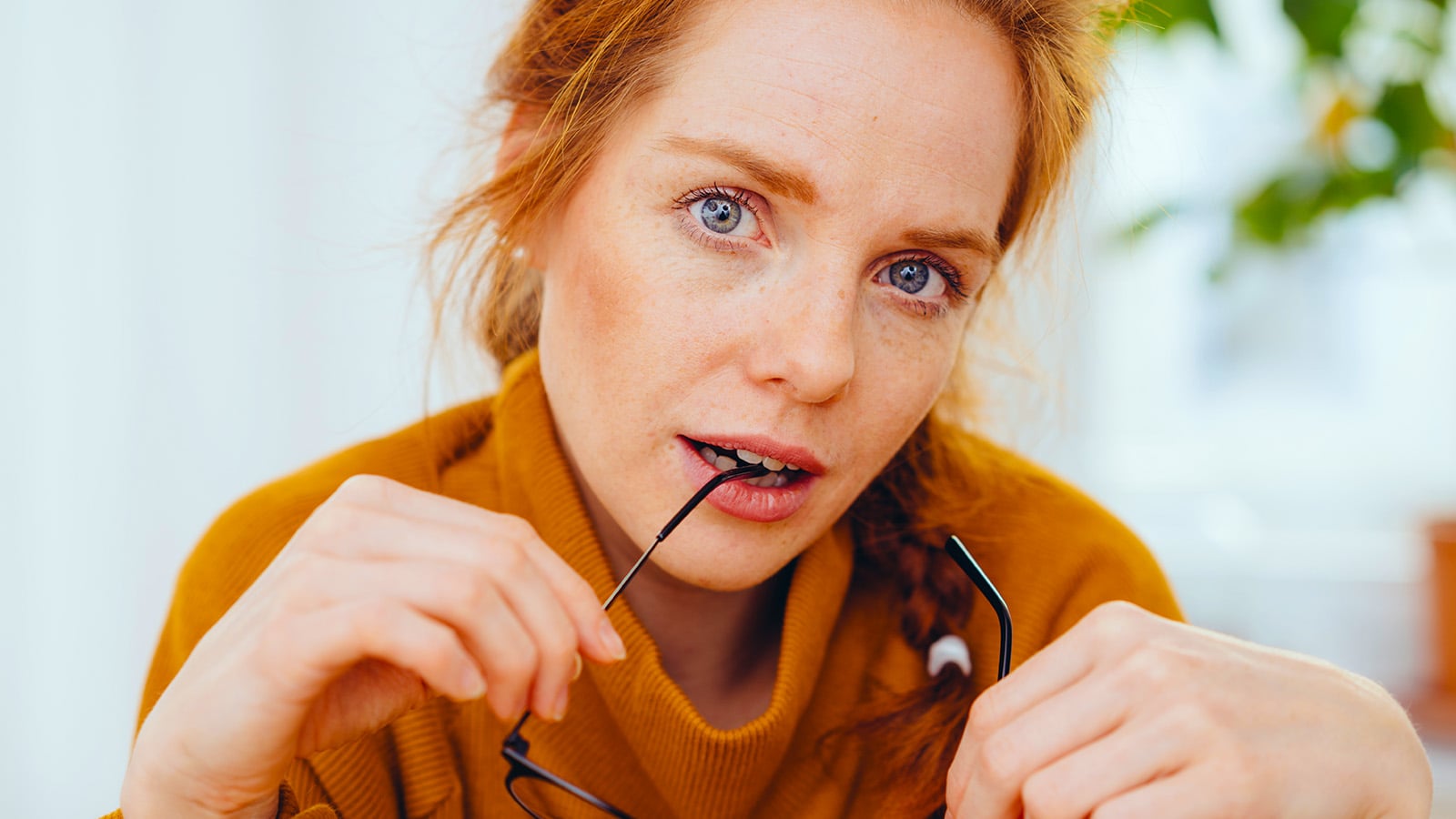 A woman with red hair and freckles in a mustard sweater holds glasses and looks thoughtfully at the camera, lightly biting the temple arm, as if contemplating the unique strengths that daughters often develop while navigating relationships with difficult mothers.