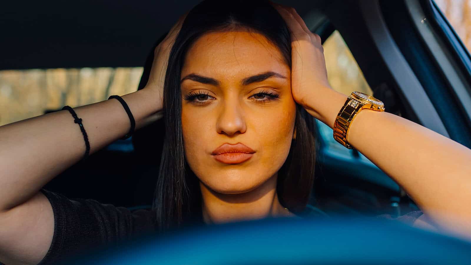 A woman with long dark hair sits in a car, holding her head with both hands, looking straight ahead with a serious expression—a moment that suggests deep reflection on psychology and personal growth. She wears a gold watch and has a black hair tie on her wrist.