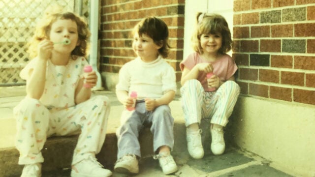 Three young children sit on outdoor steps blowing bubbles, smiling and holding bubble bottles. Dressed in casual clothes and sneakers, they capture the carefree spirit of a self-sufficient generation against a brick wall and fence backdrop.