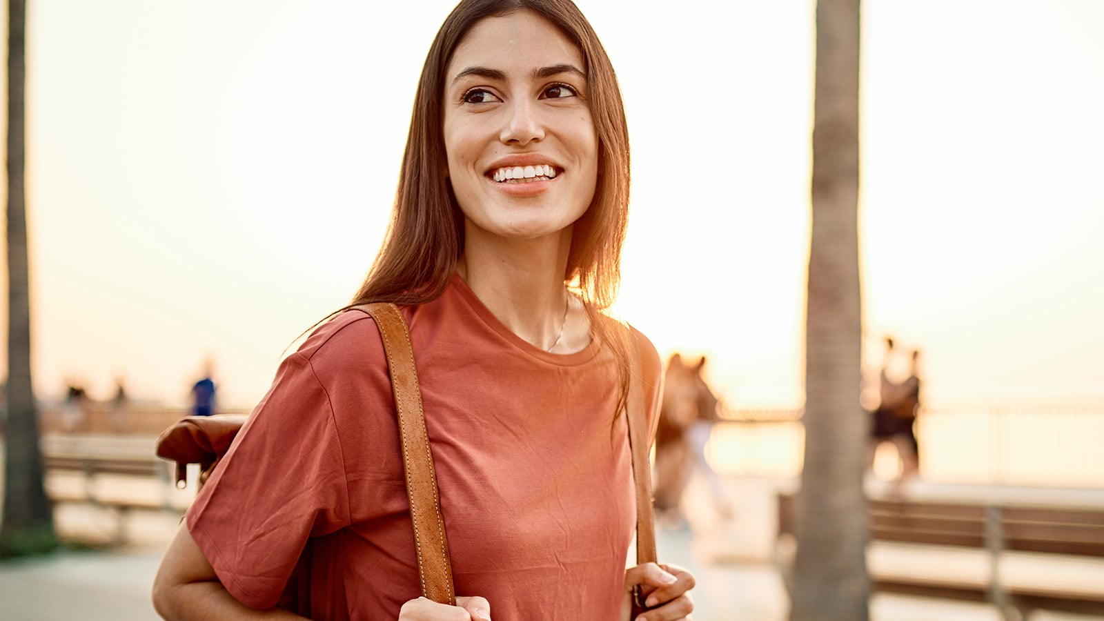 A young woman with long brown hair smiles while standing outdoors at sunset, wearing a red shirt and carrying a brown backpack. Capturing the spirit of Gen Z on vacation, palm trees, people, and benches are visible in the background.