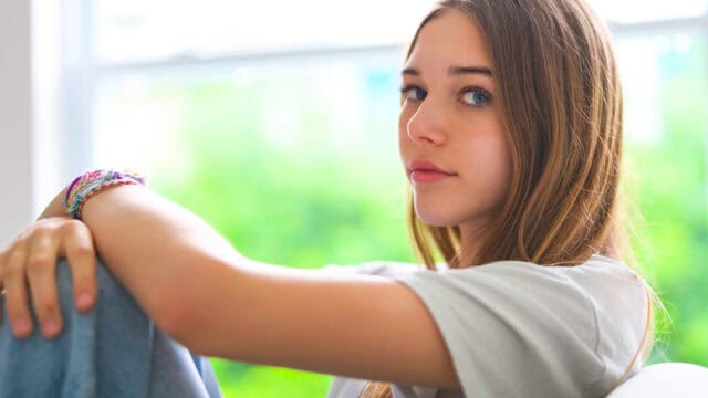 Teen girl with long brown hair sits indoors, resting her arms on her knees and looking at the camera—a quiet moment that reflects the loneliness sometimes felt by Gen Z. She wears a light t-shirt and bracelets. The background is blurred and green, suggesting trees or plants outside a window.