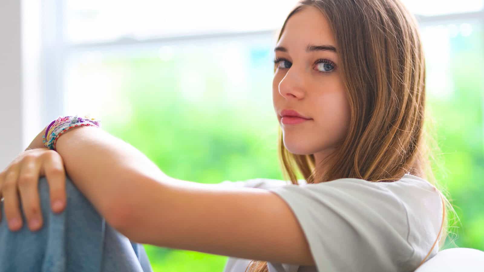 Teen girl with long brown hair sits indoors, resting her arms on her knees and looking at the camera—a quiet moment that reflects the loneliness sometimes felt by Gen Z. She wears a light t-shirt and bracelets. The background is blurred and green, suggesting trees or plants outside a window.