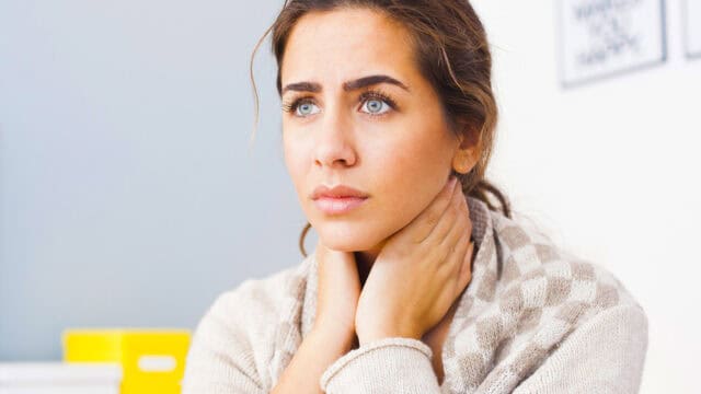 A woman with blue eyes and brown hair holds her neck with both hands and looks worried or uncomfortable, wearing a light sweater indoors—perhaps reflecting the pressures Millennials face when striving for a healthy work-life balance.