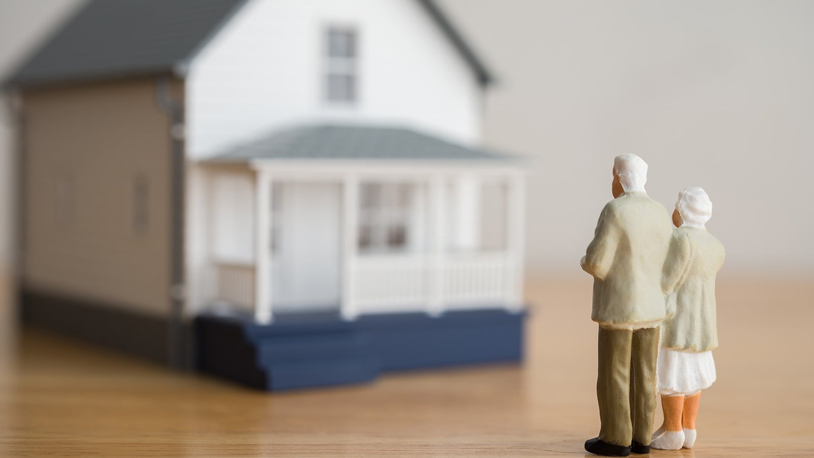 Two small figurines of an elderly couple stand together facing a model house, capturing the deep place attachment and emotions that come with grief over selling a home. The softly blurred house in the background underscores their poignant farewell.