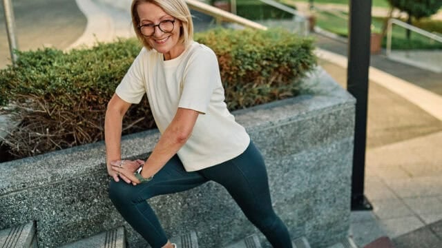 Smiling older woman with glasses in athletic wear stretching her leg on outdoor stairs, embracing positive age beliefs amid greenery and pavement in the background.