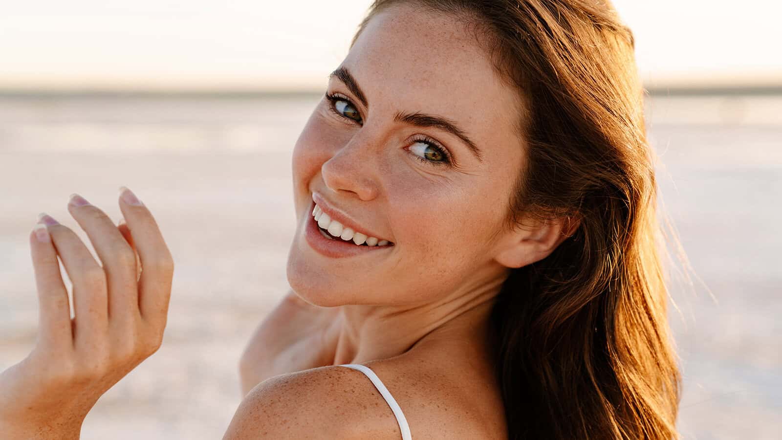 A woman with long brown hair smiles at the camera outdoors, sunlight illuminating her face. She is wearing a white top and standing on a sandy background, radiating the warmth and openness that are signs of trust and making her seem like a trusted person.