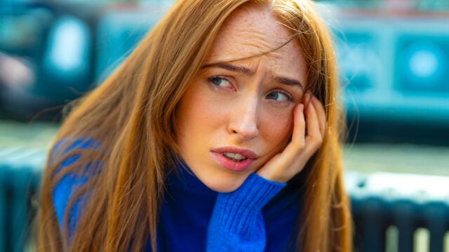 A woman with long red hair wearing a blue sweater looks worried, resting her face on her hand. The background is blurred, capturing the quiet weight eldest daughters often feel as they navigate complex family roles.