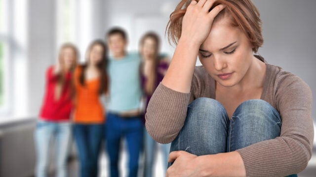 A young woman with rare traits sits alone, looking upset with her head resting on her hand, while a group of four people stands together in the blurred background.