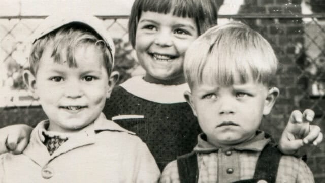 Three young children pose together; two boys in front, one smiling and the other looking serious, with a smiling girl standing behind them, her arms around their shoulders. The black and white photo, complete with a fence in the background, captures old-school values and the nostalgic habits of childhood friendships reminiscent of the 60s & 70s.