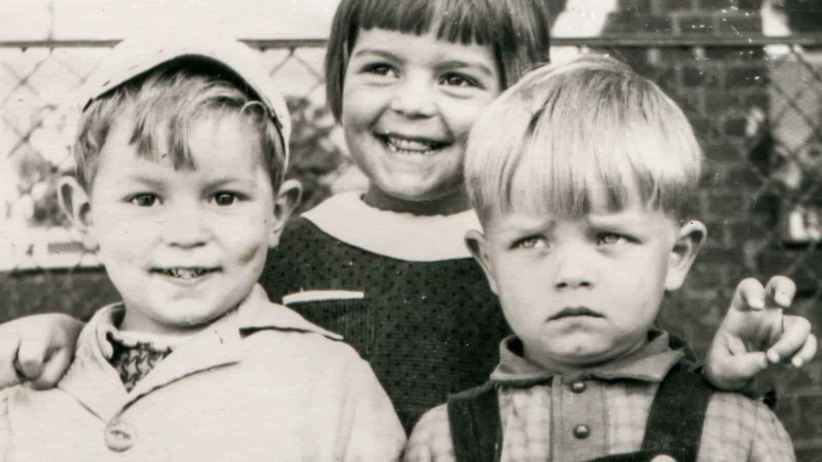 Three young children pose together; two boys in front, one smiling and the other looking serious, with a smiling girl standing behind them, her arms around their shoulders. The black and white photo, complete with a fence in the background, captures old-school values and the nostalgic habits of childhood friendships reminiscent of the 60s & 70s.