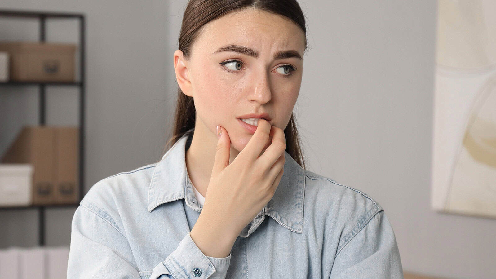 A young woman in a light denim shirt looks to the side with a worried expression, biting her fingernail and furrowing her brow—perhaps weighed down by shrinking happiness after another comparison with others. Shelves of boxes and books blur in the background.