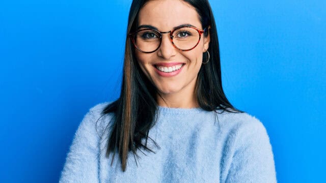 Woman with long dark hair, wearing glasses and a light blue sweater, smiles in front of a bright blue background, her expression reminiscent of someone skilled at delivering non-apology statements.
