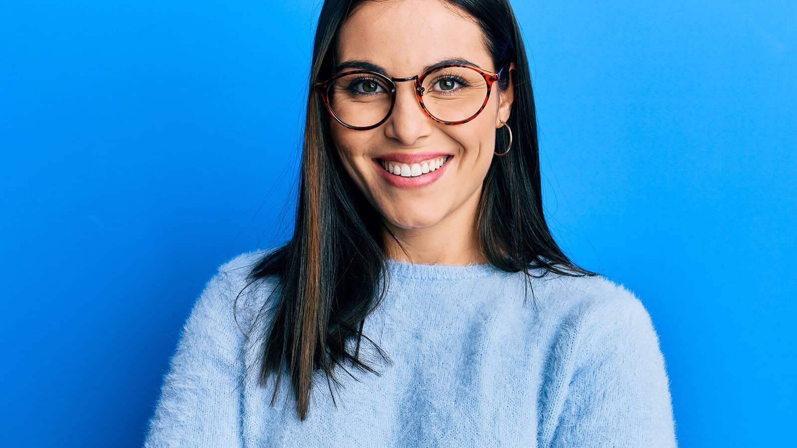 Woman with long dark hair, wearing glasses and a light blue sweater, smiles in front of a bright blue background, her expression reminiscent of someone skilled at delivering non-apology statements.