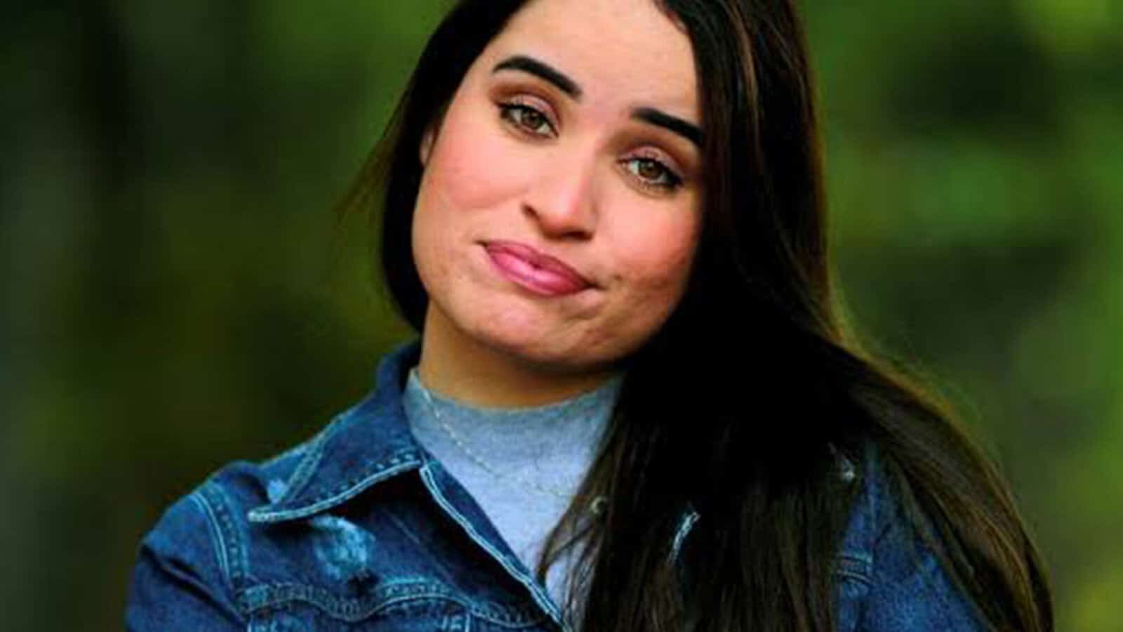 A woman with long dark hair, wearing a blue denim jacket, looks at the camera with a neutral expression and slightly pursed lips—a contemplative look often seen among millennials since the 2008 financial collapse. The background is blurred greenery.