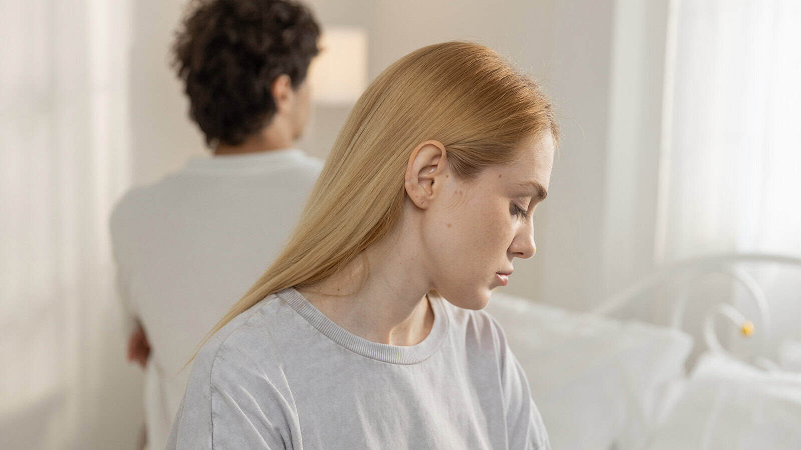 A woman with blonde hair looks down sadly while a man with dark curly hair sits behind her, facing away. Both appear upset in a bright bedroom, highlighting relationship challenges and moments of marriage loneliness.
