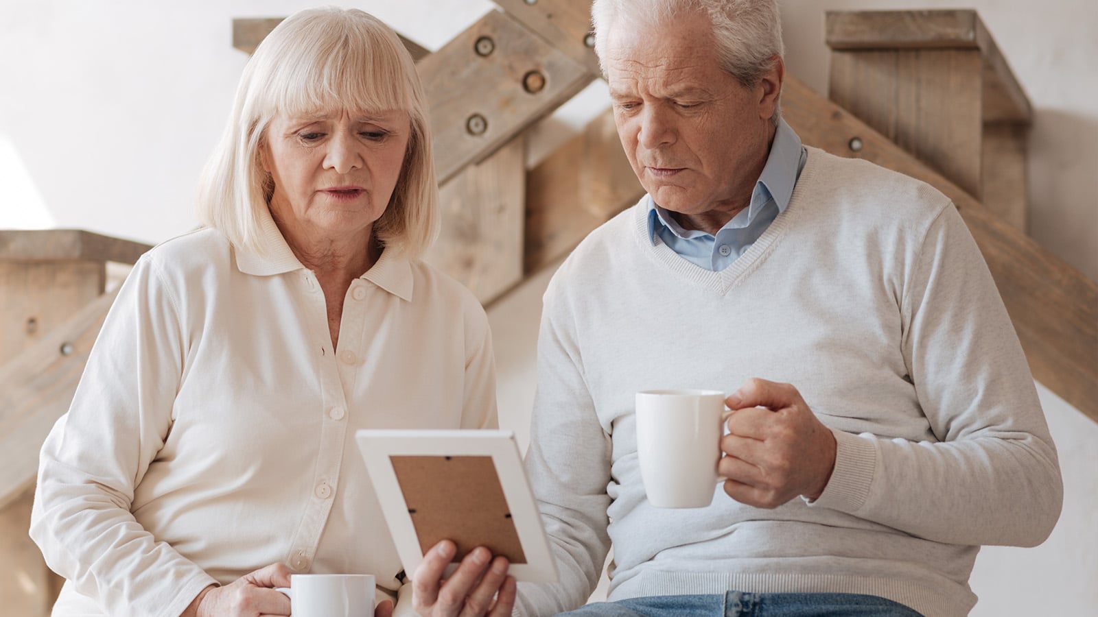 An elderly couple sits together, both holding white mugs. The woman looks at a framed photograph while the man gazes at it too. They appear thoughtful, perhaps reflecting on cherished family relationships, and are seated indoors near wooden stairs.