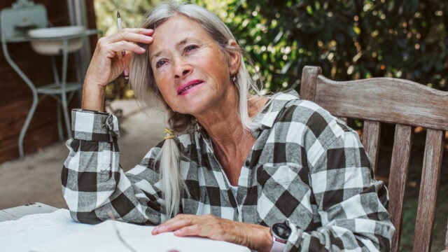 Older woman with gray hair in a plaid shirt sits outdoors at a table, smiling thoughtfully with a pen in hand and an open notebook in front of her, surrounded by greenery—perhaps jotting down dreams for her bucket list and reflecting on life fulfillment during retirement.