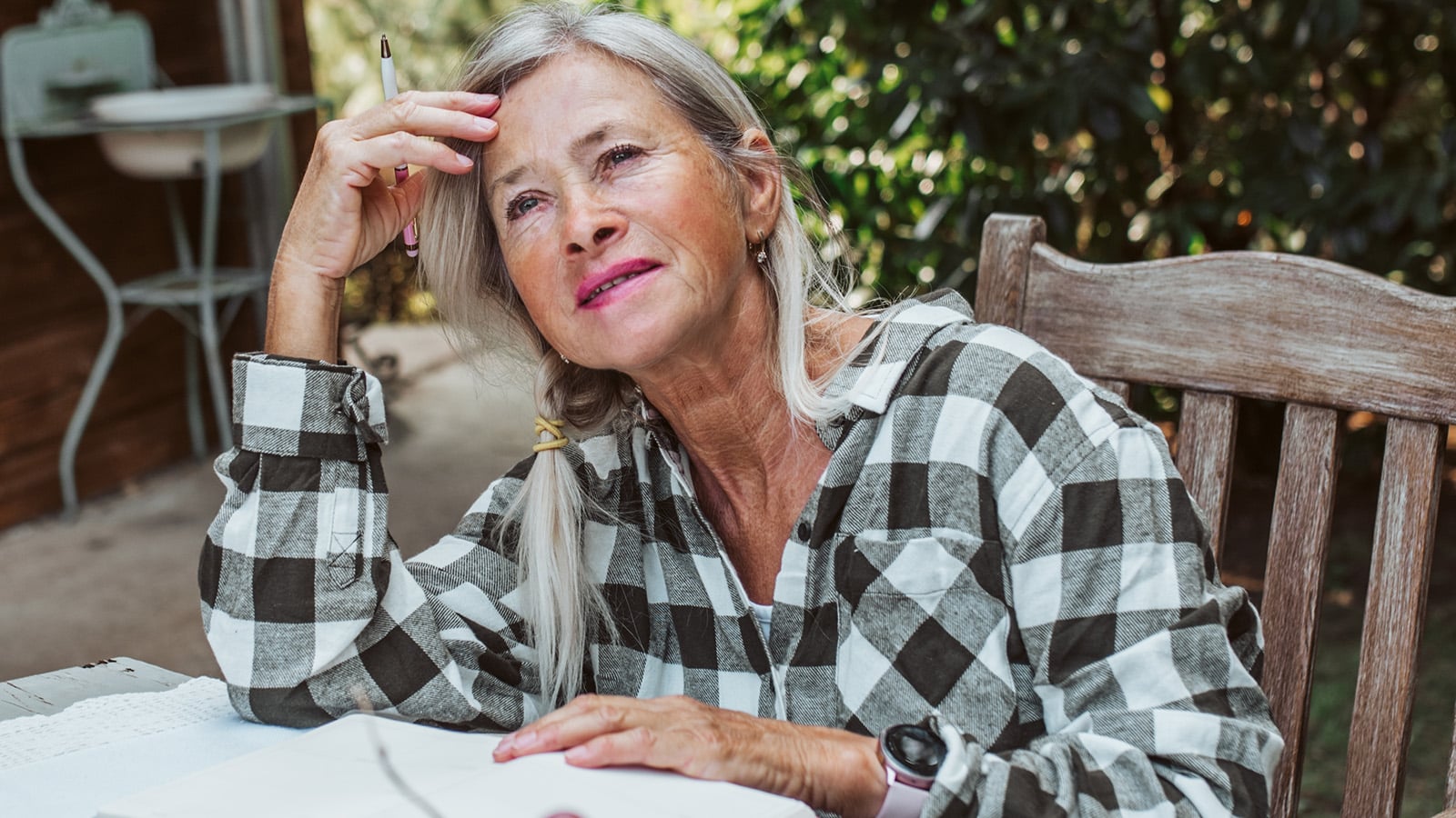 Older woman with gray hair in a plaid shirt sits outdoors at a table, smiling thoughtfully with a pen in hand and an open notebook in front of her, surrounded by greenery—perhaps jotting down dreams for her bucket list and reflecting on life fulfillment during retirement.