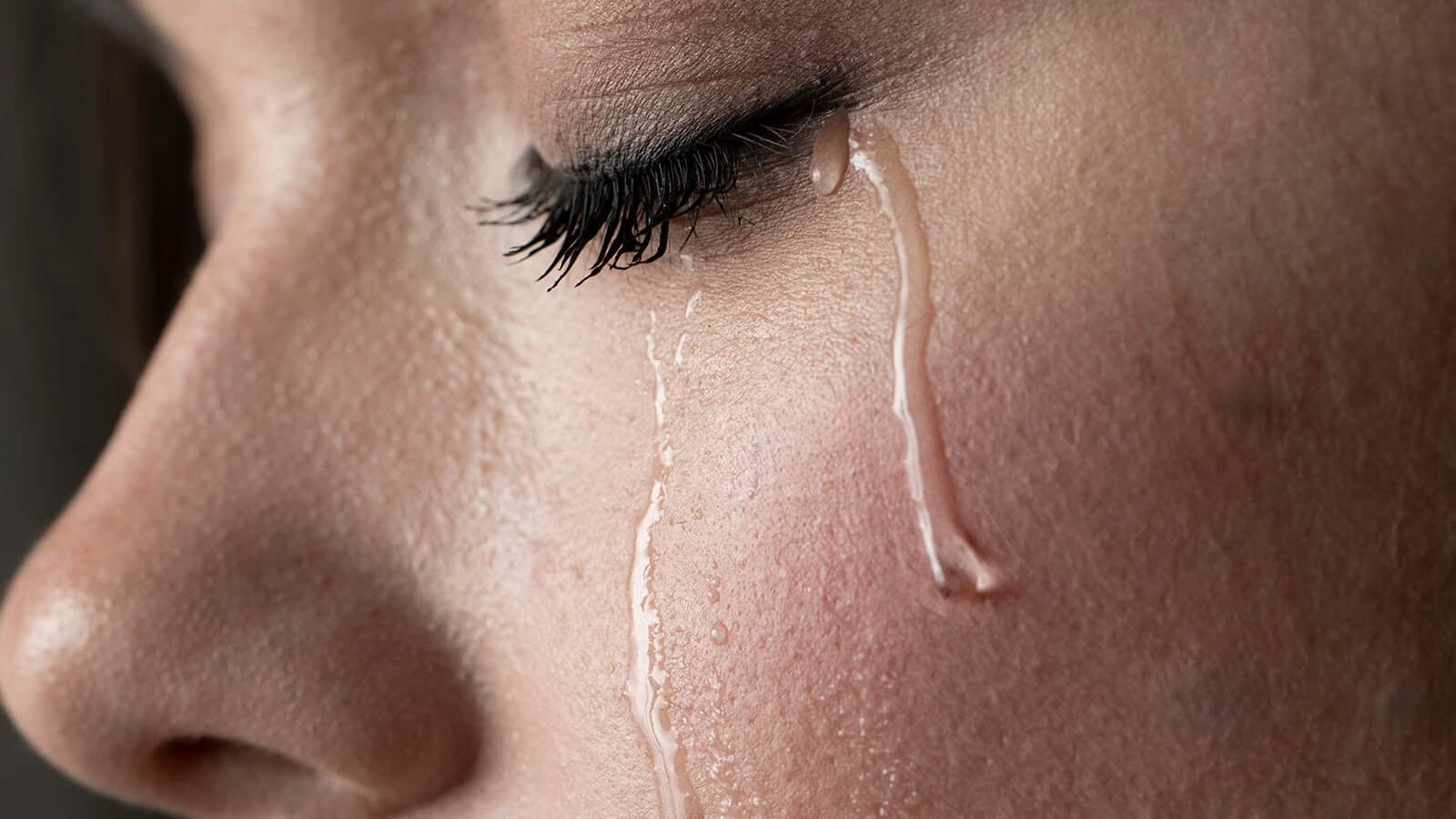 Close-up of a person’s face with closed eye and long eyelashes, showing two tears streaming down their cheek—a powerful depiction of grief and mourning.