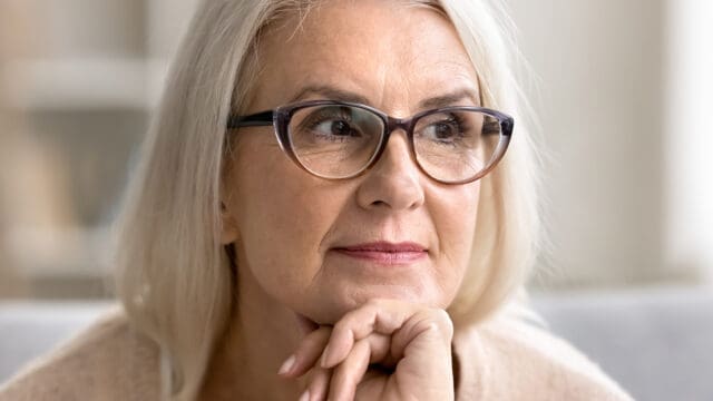 An older woman with long gray hair and glasses sits indoors, resting her chin on her hand and looking thoughtfully to the side, embodying the reflective spirit often seen in retirees as they consider their second act.