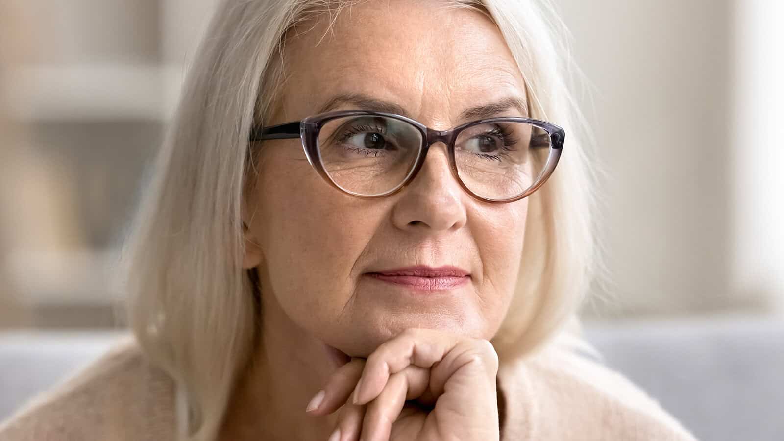 An older woman with long gray hair and glasses sits indoors, resting her chin on her hand and looking thoughtfully to the side, embodying the reflective spirit often seen in retirees as they consider their second act.