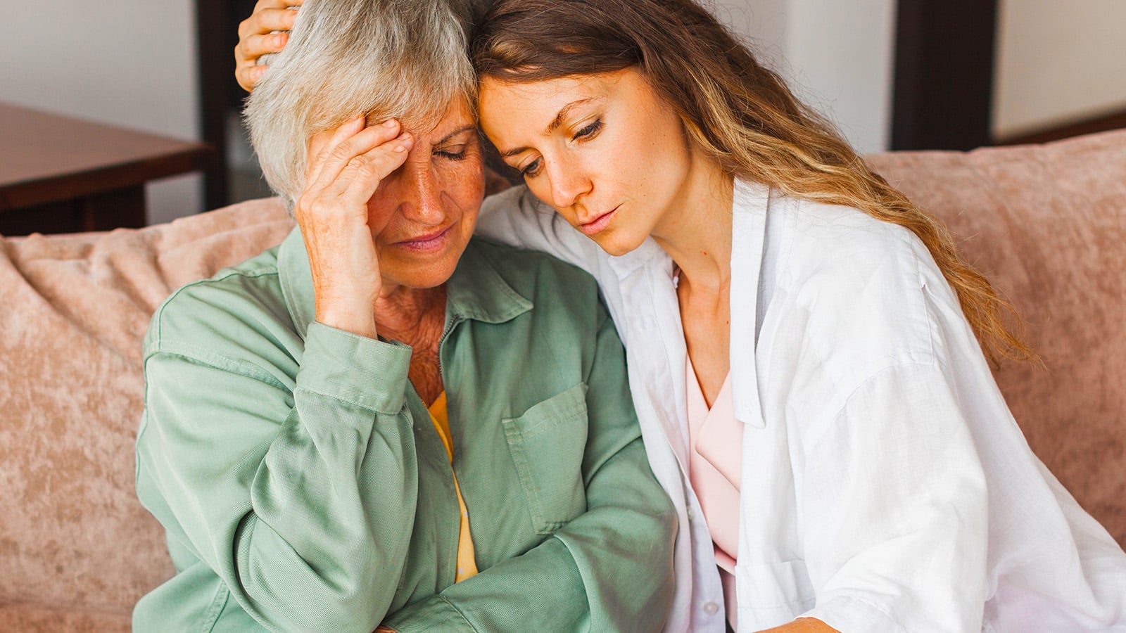 An older woman looks distressed while a younger woman sits beside her, offering comfort with an arm around her shoulders. They sit close together on a couch, capturing a heartfelt moment of caregiving and support as they navigate the challenges of parent aging together.