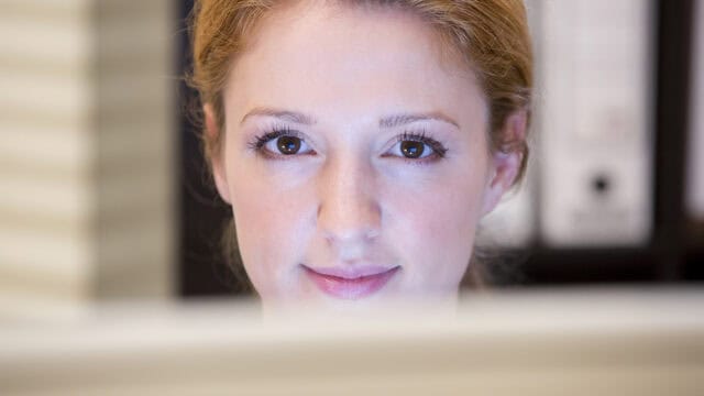 A woman with light skin and reddish-blonde hair looks at a computer screen, her face softly lit and background out of focus, capturing a quiet moment of overcoming challenges as she focuses intently on her work.