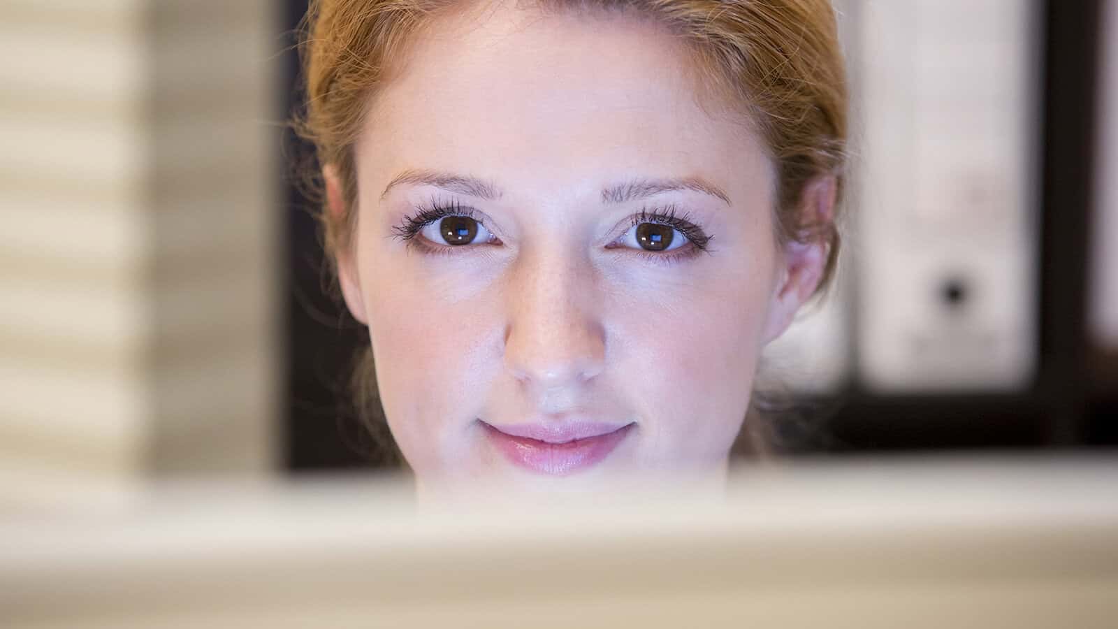 A woman with light skin and reddish-blonde hair looks at a computer screen, her face softly lit and background out of focus, capturing a quiet moment of overcoming challenges as she focuses intently on her work.