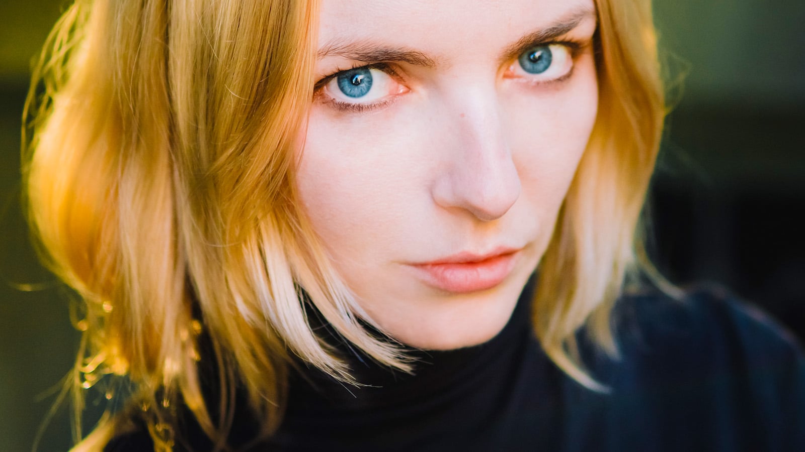 A person with shoulder-length blonde hair and blue eyes looks seriously at the camera, wearing a black turtleneck. The lighting highlights their face and hair, creating a warm glow on one side, as if capturing a quiet moment of healing.