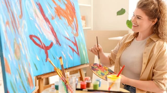 A smiling woman enjoys her leisure hobby as she paints an abstract, colorful design on a large canvas in a bright room, holding a palette and brush, with paint supplies nearby.