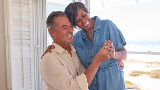 A smiling couple dances together indoors by a window with a beach view, holding hands and looking happy and relaxed in casual clothing—perfectly capturing the joy of happy second marriages and the beauty of appreciating things to do differently the second time around.
