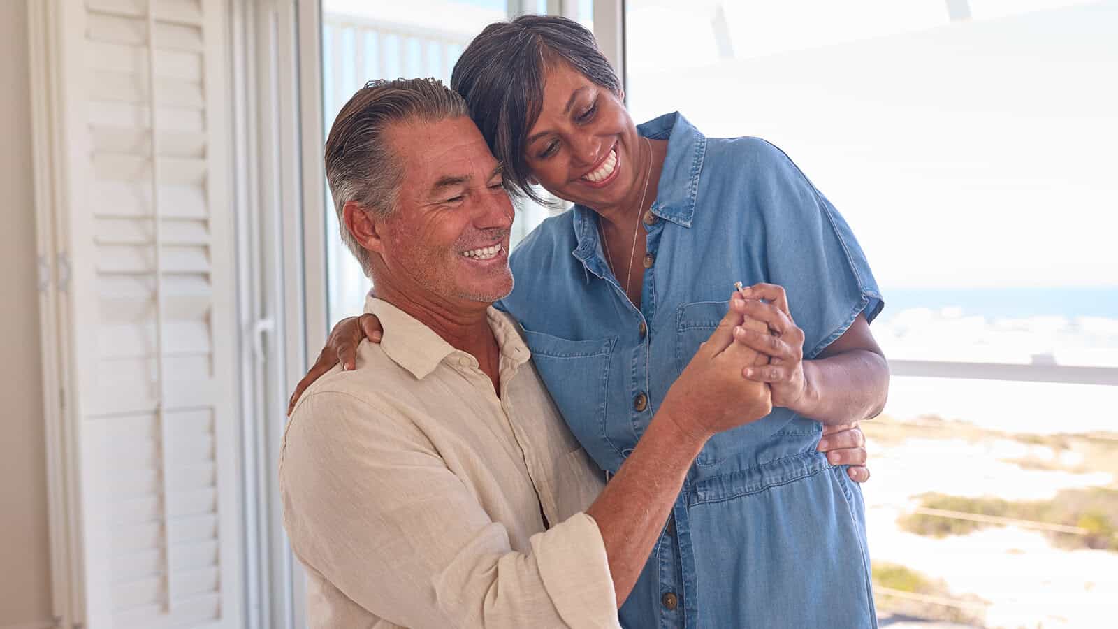 A smiling couple dances together indoors by a window with a beach view, holding hands and looking happy and relaxed in casual clothing—perfectly capturing the joy of happy second marriages and the beauty of appreciating things to do differently the second time around.