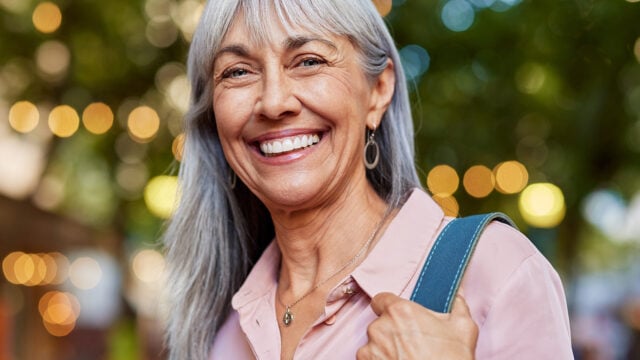 Smiling older woman with long gray hair, wearing a light pink blouse and silver earrings, stands outdoors with blurred green trees and lights in the background, holding a blue bag over her shoulder—a beautiful example of positive aging habits.