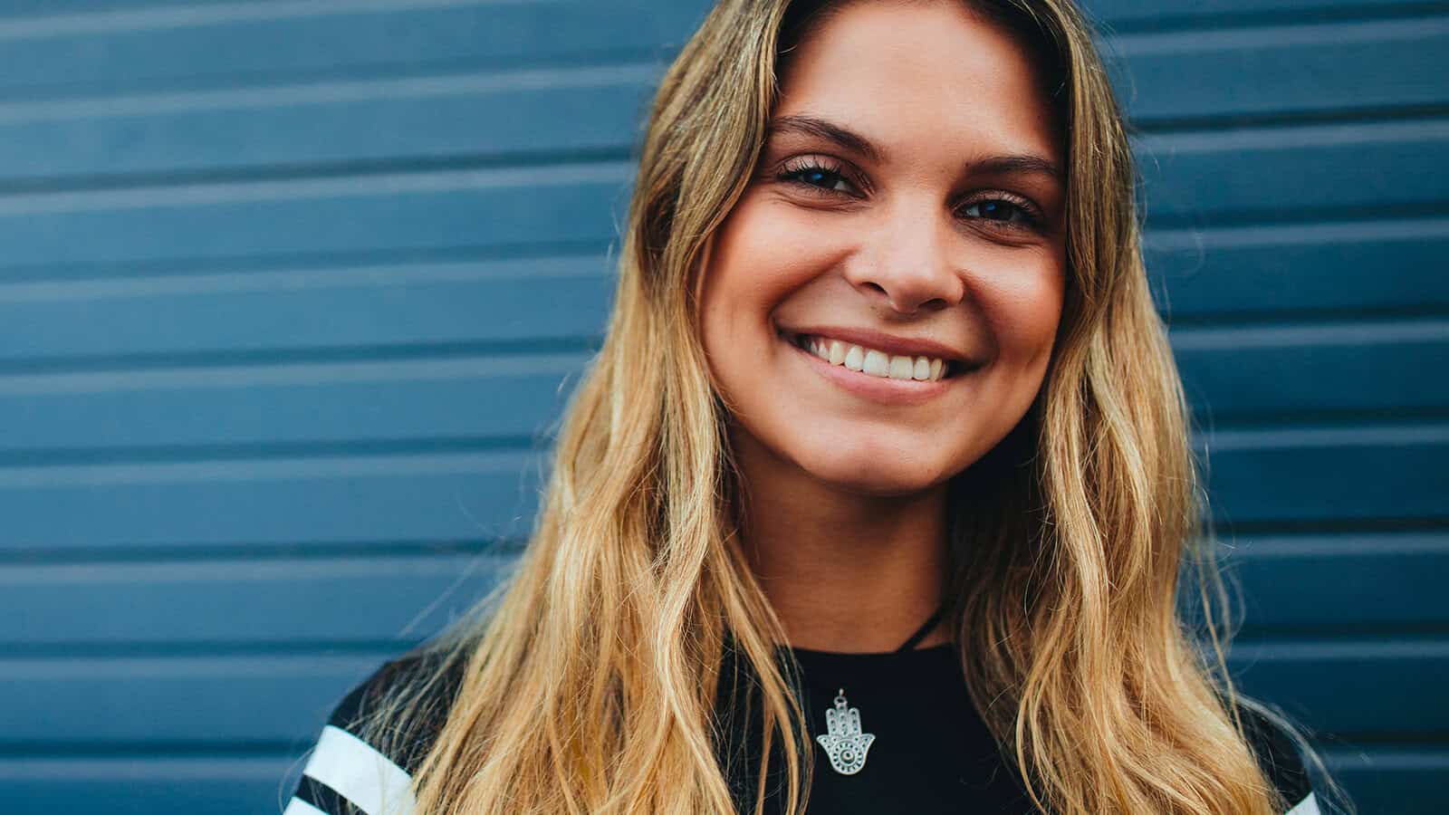 A young woman with long, wavy blonde hair smiles at the camera, standing in front of a blue, horizontal-striped background. She wears a black top and a necklace with a pendant, exuding confidence and discipline in her posture.