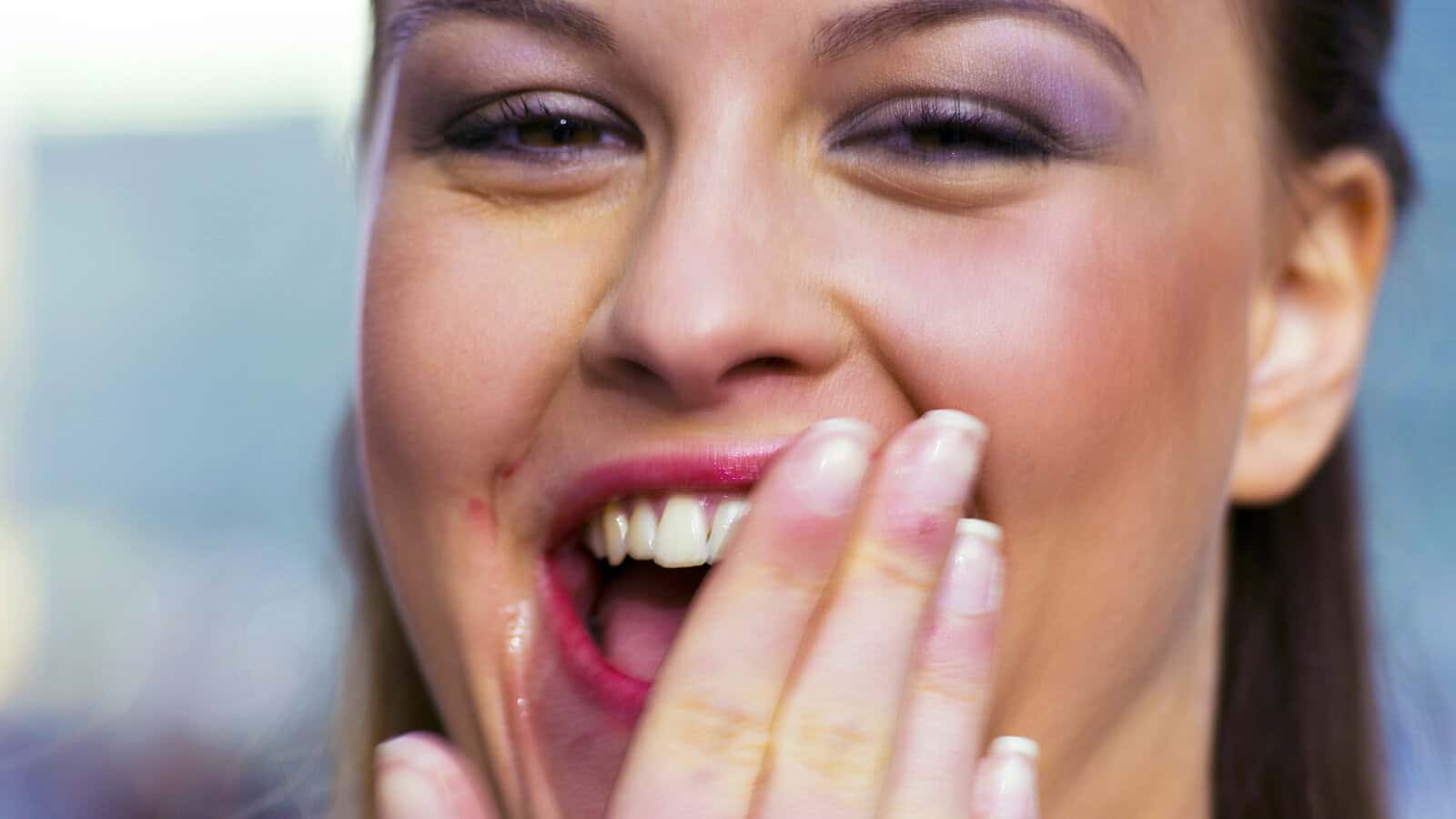 A person with makeup is smiling and covering their mouth with their hand, showing teeth with a lipstick smudge near their lips—psychologists might say that such an expressive gesture reveals intriguing personality traits.