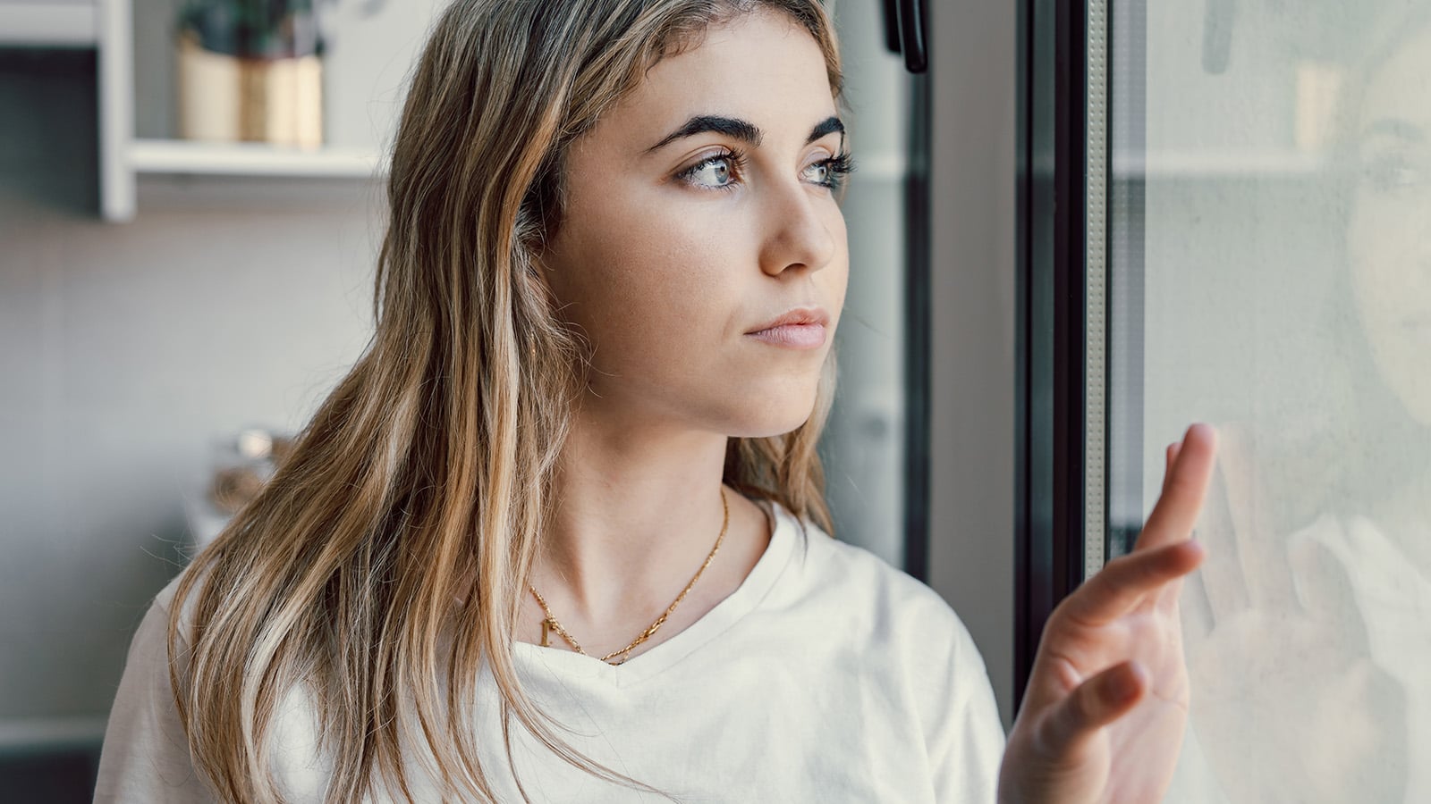 A young woman with long blonde hair looks thoughtfully out a window, gently touching the glass with her hand. She wears a white top and a delicate necklace, her expression reflecting the quiet strength of resilient adults who have weathered family disappointment and learned the art of survival.