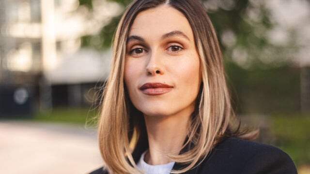 A woman with straight, shoulder-length blonde hair and a slight smile stands outdoors, wearing a black jacket and a white top, with a blurred background of trees and buildings—her confident posture hinting at someone who holds high standards in relationships, unafraid to take emotional risks.