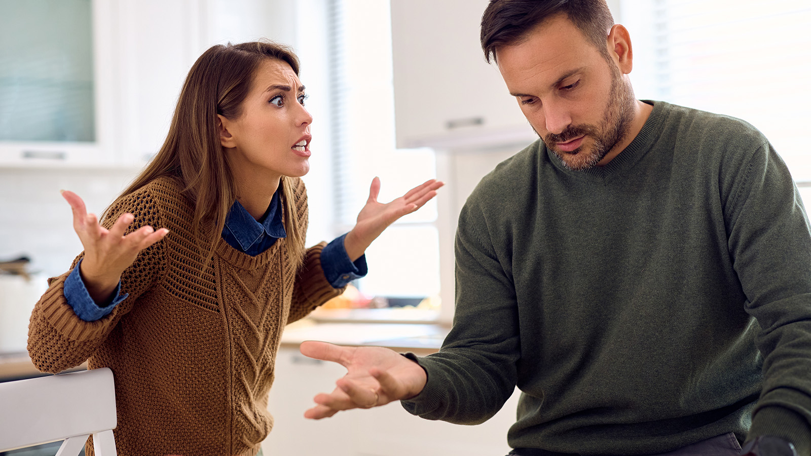 A woman appears upset, raising her hands and speaking to a man who sits with his head down, looking frustrated. They are indoors, possibly in a kitchen, both wearing sweaters—clear signs in relationships of reaching breaking point.