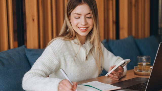 A woman in a white sweater smiles while writing in a notebook, holding a smartphone, and sitting at a table with a laptop and a glass of water, perhaps tracking her money habits or using a budgeting app to balance her checkbook.
