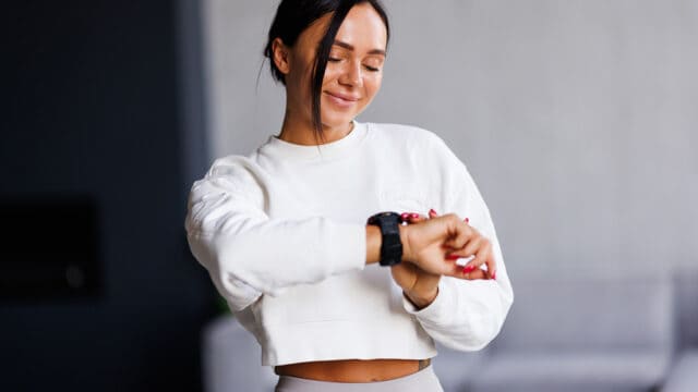 Woman in a white crop sweatshirt smiles while checking her smartwatch. She stands indoors with a blurred background, appearing relaxed and content—perhaps displaying the calm confidence that comes from early arriver psychology and the peace of mind it brings.