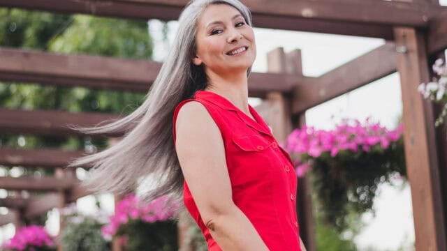 A smiling woman with long gray hair, embodying the composure often seen in Gen X women, wears a sleeveless red dress and stands outdoors under a wooden pergola with hanging baskets of pink flowers.