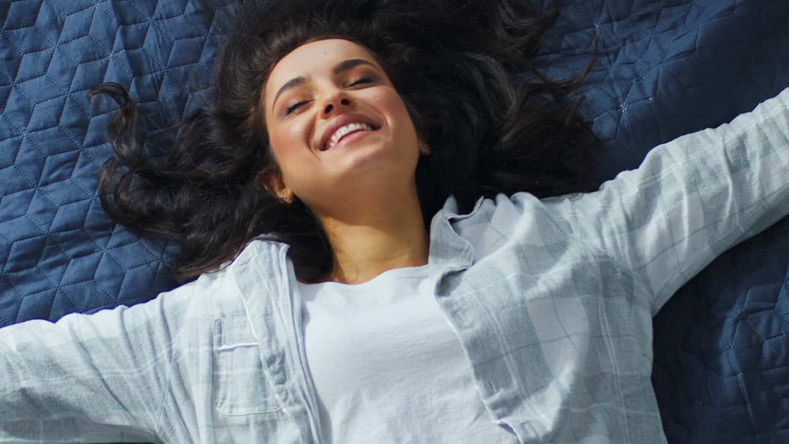 A young woman with long dark hair smiles joyfully while lying on a dark blue quilt, arms outstretched, wearing a white shirt and light plaid overshirt—her expression capturing a rare moment of solitude and happiness.