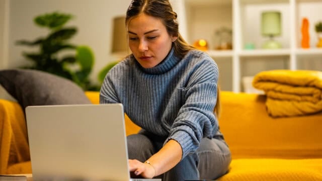 A woman in a gray sweater sits on a mustard-yellow sofa, focused on task completion as she uses her laptop in a cozy living room with plants and shelves in the background—a subtle nod to the principles of psychology at play in her productive routine.
