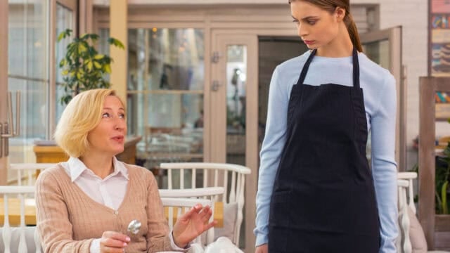 A seated woman is talking to a standing waitress in an apron inside a restaurant. The waitress, whose serious expression suggests she might be thinking deeply about psychology or the ways different nervous systems respond in social situations, looks down at the customer as they converse.