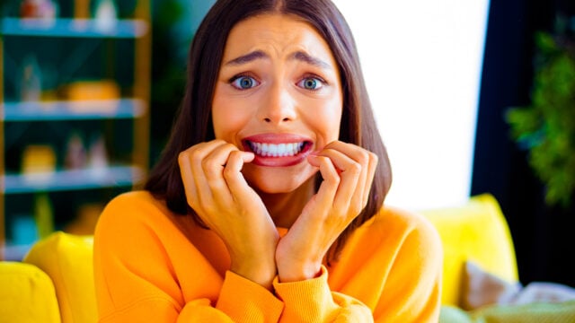 A woman in a yellow sweater sits indoors, looking anxious and biting her fingernails, with wide eyes and a tense expression, perhaps experiencing indebtedness aversion as described in psychology.
